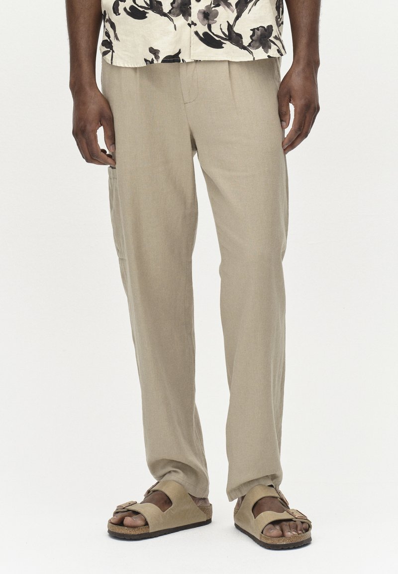 Pantalons en lin beige clair avec une coupe décontractée, dotés d'une poche latérale. Associés à des sandales beiges confortables. Chemise à manches courtes à motifs floraux visible.