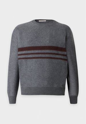 Homme portant un pull gris à rayures marron, un pantalon ample marron foncé et des bottes rouge foncé avec des lacets rouges, debout devant un fond uni.