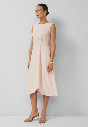 Vrouw draagt een mouwloze, lichtroze midi-jurk met geplooide voorkant, gouden armband, oorringen en beige sandalen met hak, staand tegen een effen muur.