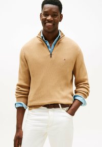 Uomo sorridente che indossa un maglione color cannella a coste con mezza zip sopra una camicia blu con le maniche rimboccate, pantaloni bianchi e cintura marrone, in piedi con una mano in tasca.
