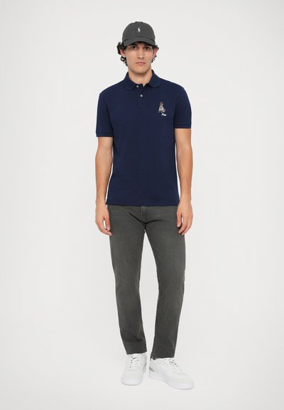 Camisa polo azul-marinho de manga curta com design bordado, combinada com jeans cinzentos e ténis brancos. Homem está de pé contra um fundo simples.