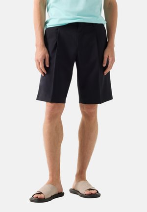 Homme portant un short noir mi-cuisse et des sandales beige à enfiler, debout devant un fond clair uni.