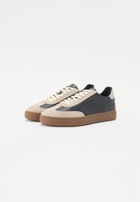 Beige og marineblå sneakers laget av semsket skinn og lær, med en kraftig gummisåle, flate snører og merkede detaljer.