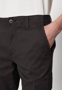 Ciemnobeżowe spodnie o gładkiej, matowej fakturze, z zapięciem na guzik z kontrastowym akcentem oraz boczną kieszenią dla funkcjonalności.