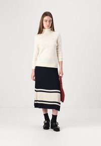 Kremowy sweter z golfem noszony z granatową plisowaną spódnicą w beżowe paski. Czarne buty na blokowym obcasie i czerwona torebka. Minimalistyczny design.
