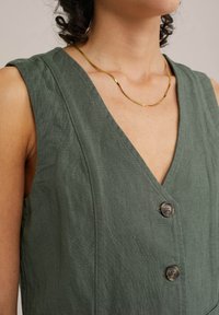 Zelené lněné šaty s hlubokým výstřihem do V, bez rukávů a se dvěma knoflíky vpředu. Obsahují texturovanou látku pro přidaný detail.