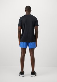 Sort kortærmet t-shirt, blå sportsshorts og sorte sneakers. Mand står med ryggen til en ensfarvet hvid baggrund.