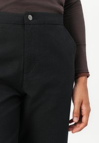 Close-up of a person wearing black pants with a button labeled "RVCA" and a brown long-sleeve top, hand relaxed by the side.