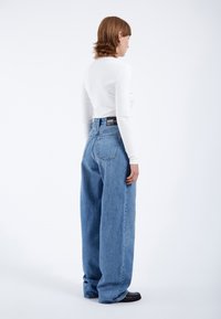 Vrouw met schouderlang haar, gekleed in een wit topje met lange mouwen, een high-waist brede blauwe spijkerbroek en zwarte schoenen, staand tegen een witte achtergrond.