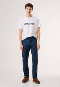 Bílé bavlněné tričko s logem „PEPE JEANS“ v námořnické modři a červené barvě, doplněné tmavě modrými kalhotami a hnědými semišovými šněrovacími botami.