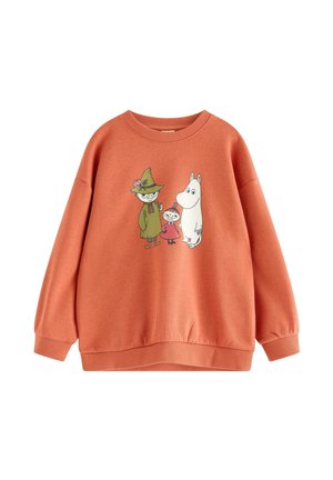 Ung dreng smilende og lænende let fremad, iført en orange sweatshirt med tegneseriefigurer, blå jeans og beige sneakers.