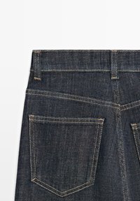 Mörkblå denimjeans med hög midja, två bakfickor med kontrasterande sömmar och en slät yta. Kanterna är prydligt avslutade.