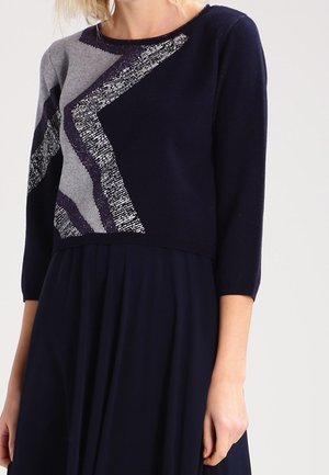 Kobieta w granatowej sukience z rękawami do połowy ramienia i abstrakcyjnym wzorem zygzaków w odcieniach szarości i bieli na górnej części sukienki.