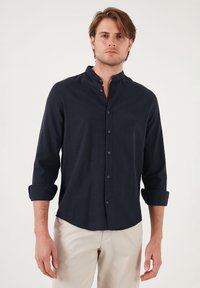Camisa azul marino de botones con cuello mao, mangas largas arremangadas en los puños y una tela texturizada. Presenta un sutil patrón tejido.