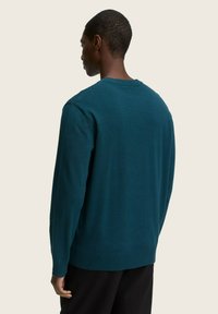 Türkis gestrickter Pullover mit langen Ärmeln, gerippten Bündchen und Kragen. Glatte Textur, lässige Passform, sichtbare Nahtdetails, getragen über schwarzen Hosen.
