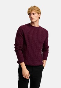 Bordeaux kabelstickad tröja med rund halsringning och ribbade ärmslut, matchad med svarta byxor. Strukturerad tyg med upphöjda mönster.