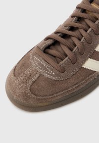 Brun semsket joggesko med hvite striper, rund tå, teksturert overdel, snøring og gummisåle med tydelige sømdetaljer.