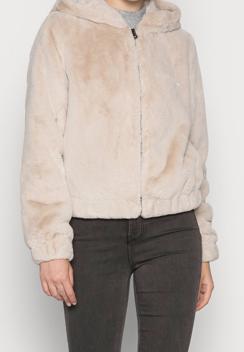 Lys beige faux fur jakke med hætte, lynlås foran og elastiske manchetter. Blød tekstur og let cropped design.