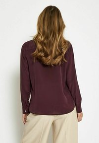 Blusa borgogna a maniche lunghe con una texture liscia, orlo arrotondato e polsini con bottoni. Presenta una leggera arricciatura sulle spalle.