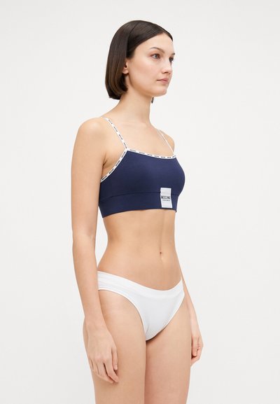 Top corto blu navy con sottili spalline bianche e accento con logo; abbinato a un brief bianco a vita bassa; texture liscia e design aderente.