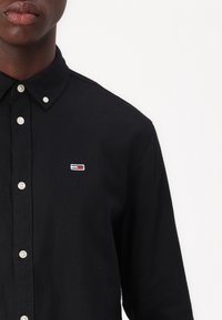 Czarna koszula z kołnierzykiem na guziki. Ma białe guziki oraz małe logo w kolorach czerwonym, białym i niebieskim na lewej piersi. Wykonana z bawełny.