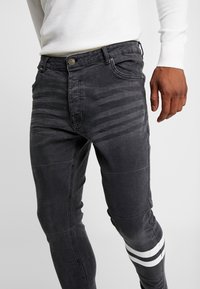 Slim-fit jeans in dark gray denim with subtle vertical stripes, featuring a white stripe detail on the lower leg and metal button hardware.