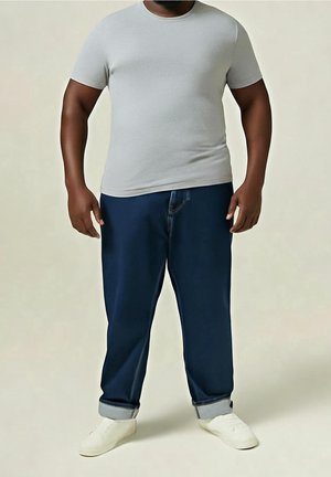 Mężczyzna stojący w jasnoszarej przylegającej koszulce, ciemnoniebieskich podwiniętych dżinsach i białych trampkach na jednolitym tle.