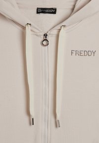 Felpa beige con zip, realizzata in tessuto morbido, con lacci bianchi, hardware argentato e la scritta "FREDDY" in strass sul davanti.