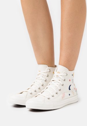 Personne portant des baskets hautes en toile blanche avec des broderies colorées de motifs célestes et végétaux, debout sur un fond blanc uni.