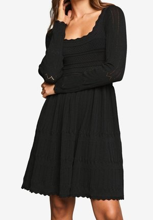 Femme portant une robe en maille texturée noire à manches longues avec un col carré et des bords festonnés.