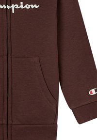 Sudadera con cremallera marrón hecha de una mezcla de algodón. Presenta un bolsillo frontal, puños acanalados y un logo blanco de "Champion" en el pecho.