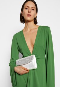 Zielona, długorękawowa sukienka z głębokim dekoltem w kształcie litery V, zestawiona z srebrną metaliczną kopertówką z błyszczącą klapką. Gładka, lejąca się tkanina.
