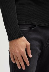 Long-sleeve black textured top with ribbed fabric, featuring a black trim at the cuffs, paired with dark jeans. Hand resting on the thigh.