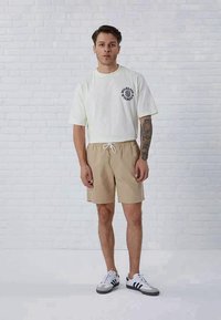 T-shirt de algodão de cor clara com estampado gráfico, combinada com calções de algodão bege com uma cintura elástica. Usado com sapatilhas brancas e cinzas.
