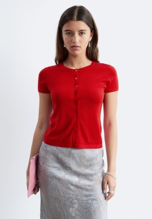 Femme aux longs cheveux bruns portant un haut rouge à manches courtes boutonné et une jupe fleurie argentée, tenant une pochette rose sur fond blanc.