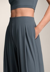 Grijze geplooide broek met een gladde textuur, voorzien van zijzakken en een hoge taille. Close-up van de stof en structuur.