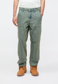 Πράσινο τζιν cargo παντελόνι με άνετη εφαρμογή, πολλαπλές πλαϊνές τσέπες και ευθεία γραμμή στο πόδι· συνδυασμένο με ταμπά εργοταξιακά μποτάκια με κορδόνια.