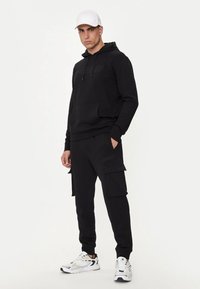 Felpa nera con logo e tasca a kangaroo, abbinata a pantaloni cargo neri con tasche laterali. Le scarpe includono sneakers bianche con dettagli.