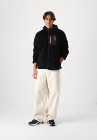 Giacca in pile nera con zip e cappuccio, con una tasca a patta nera e zipper arancione. Abbinata a pantaloni bianchi a gamba larga e sneakers scure.
