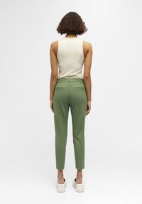 Pantalon vert fuselé avec poches arrière, taille élastique, porté avec un débardeur côtelé beige et des chaussures à plateforme blanches. Design minimaliste.