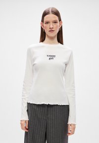Cămașă albă cu mânecă lungă, având un tiv rotunjit și manșete volan, care prezintă textul "tommy girl" brodat în negru pe piept.