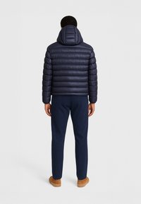 Giubbotto imbottito blu navy con cuciture orizzontali e cappuccio, abbinato a pantaloni blu navy e calzature marrone chiaro, visto da dietro.