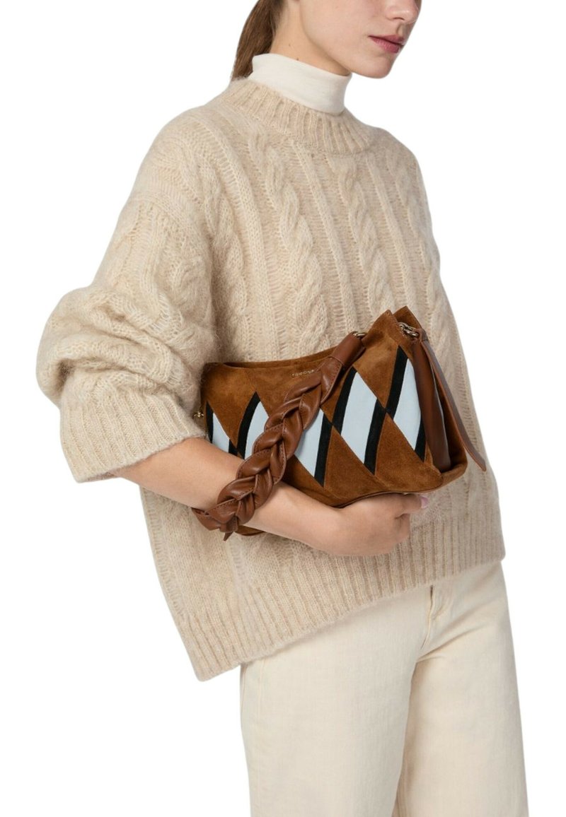 Braune Wildleder-Handtasche mit geflochtenem Lederhenkel, versehen mit diagonalen schwarz-weißen Streifen. Kombiniert mit einem beigen Strickpullover.