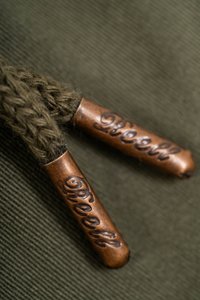 Braune Metall-Enden mit eingeprägtem Text "Reell", die an einem geflochtenen olivgrünen Cord auf einem strukturierten grünen Stoffhintergrund befestigt sind.