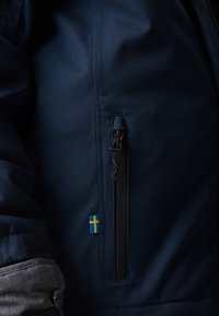 Veste bleu marine avec une poche zippée dotée d'une tirette noire et d'un petit détail de drapeau suédois sur l'ourlet. Tissu lisse, coupe cintrée.