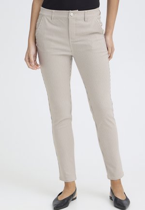 Femme portant un pantalon beige ajusté à fines rayures verticales et des ballerines noires, debout devant un fond uni.
