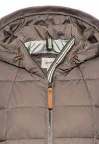 Giacca puffer imbottita marrone con cappuccio, zip frontale, fodera beige e lacci a strisce in contrasto con un accento in legno.