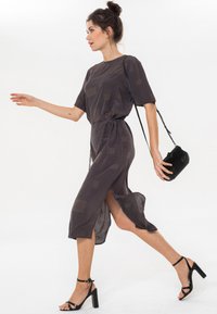 Dunkelgraues Midi-Kleid mit kurzen Ärmeln, einem Kordelzug in der Taille, geometrischen Mustern und Seitenschlitzen; kombiniert mit schwarzen, Riemchen-Absätzen und einer kleinen Tasche.