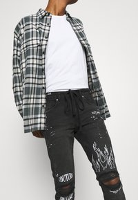 Plaid button-up shirt in green, black, and white; worn over a white t-shirt. Black jeans with paint splatter and flame graphic details.