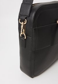 Sac en cuir noir avec une surface texturée, une forme rectangulaire, des accessoires en métal doré, une fermeture éclair et une bandoulière réglable.